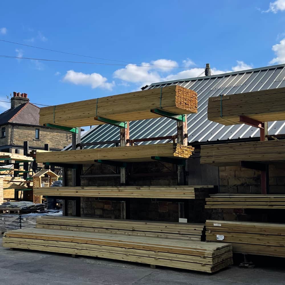 Stacks of wooden planks are neatly arranged on metal shelves outdoors, next to a building with a sloped metal roof, under a blue sky with scattered clouds.