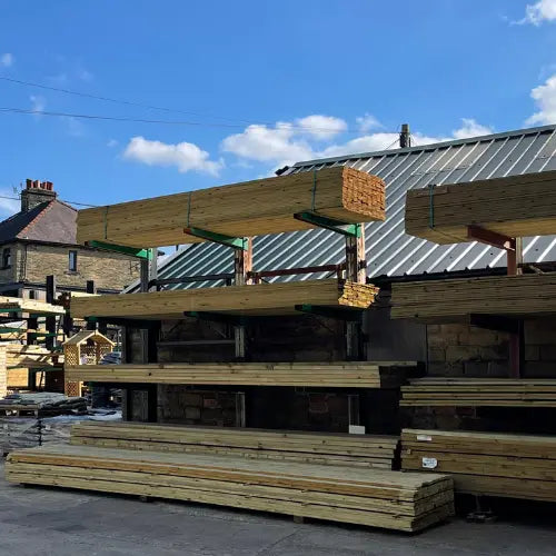 Stacks of wooden planks are neatly arranged on metal racks outdoors near a building with a gray roof, under a sunny blue sky with a few clouds.