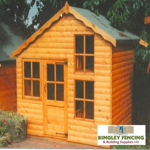 Amid lush greenery, the Upstairs Downstairs Kids Playhouse from Bingley Fencing stands with its wooden structure featuring a peaked roof, multiple windows, and a small front door. Constructed using child-friendly preservatives, it includes a sign for Bingley Fencing & Building Supplies Ltd at the bottom right.