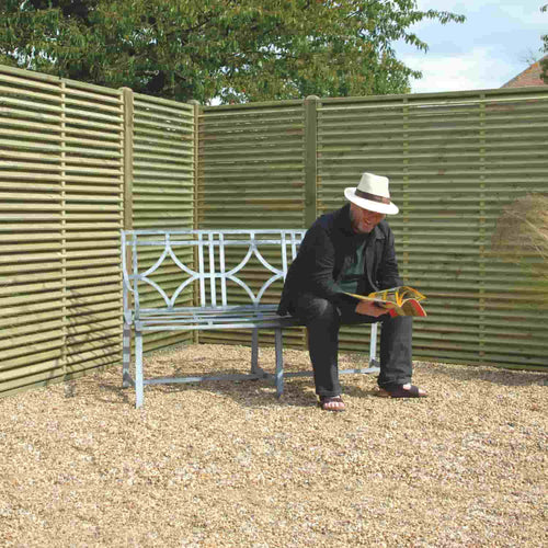 A man in a white hat and dark clothes reads a magazine on a metal bench in a gravel area bordered by KDM Reduced Venetian Fence Panels (VP180), with leafy tree branches visible above the privacy fencing.