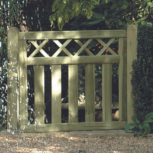 The charming Cross Top Border Gate by KDM, featuring a lattice design at the top, is framed by lush greenery. As sunlight filters through the leaves, dappled shadows play on the gravel path leading to the partially open gate, enhancing the allure of this perfect garden fence installation.