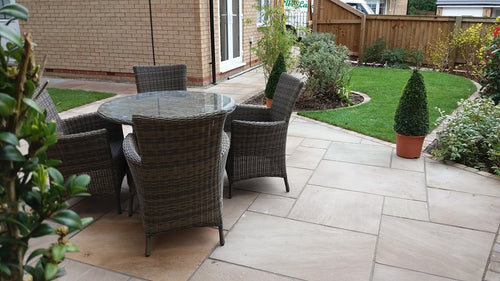 A patio featuring a set of four wicker chairs and a glass-top table on Indian Stone Paving by Bingley Fencing. The area is edged with a small green lawn, landscaped plants, and a wooden fence. A conical potted plant stands nearby, enhancing the Raj Green hues of the stones.