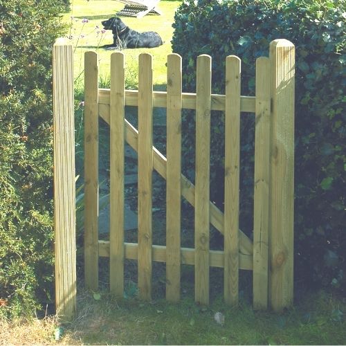 A KDM Round Top Picket Gate - RTPG120 is bordered by green hedges. Sunlight casts shadows on the grass, where a black dog lies in the background, visible through the gate slats.